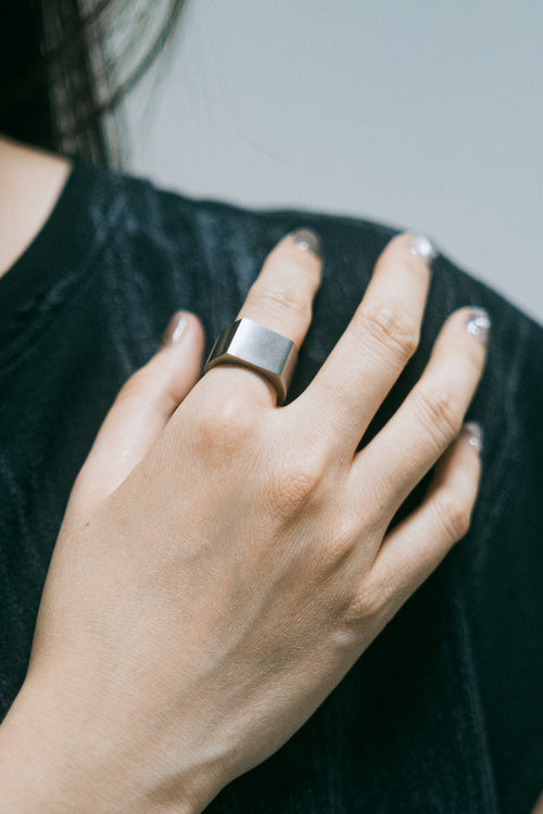 Bague Limen en acier inoxydable argenté, portée à la main posée sur l'épaule, sur fond textile sombre