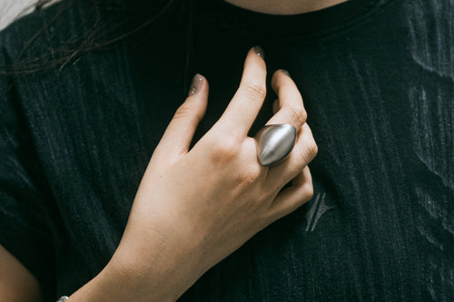 Bague Vault en acier inoxydable argenté, portée à la main posée sur la poitrine, sur fond textile sombre